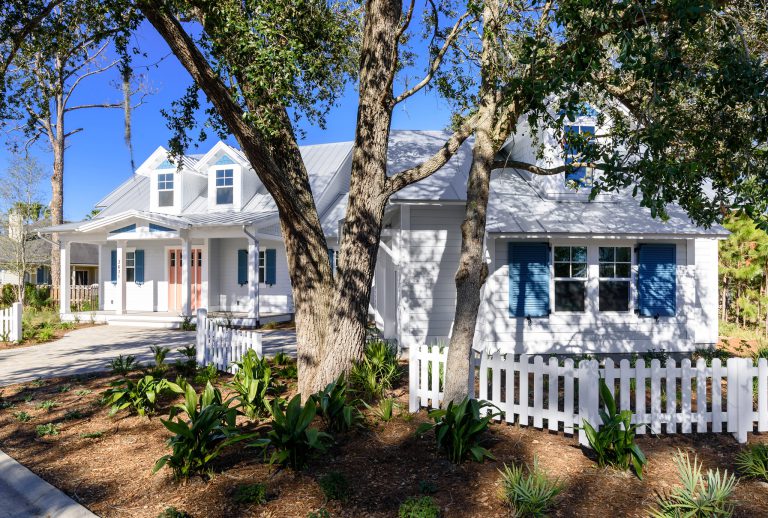 A white house with blue shutters, a metal roof, and a pink front door stands behind a white picket fence and leafy trees, with landscaped plants and a clear blue sky in the background.