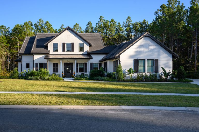 A modern two-story house with white siding, black shutters, and a dark roof, surrounded by green grass, shrubs, and trees, with a forested area in the background under a clear blue sky.