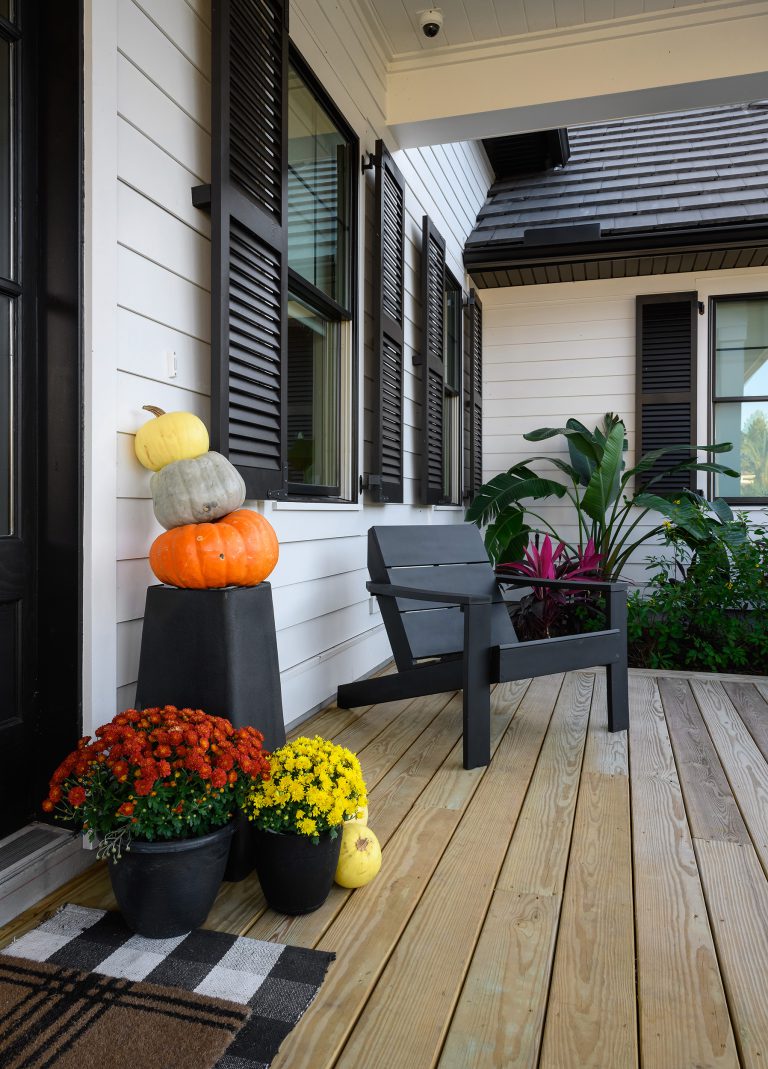 A modern porch with a black chair, stacked pumpkins on a pedestal, potted red and yellow flowers, green plants, and wood decking. The house has white siding and black shutters.