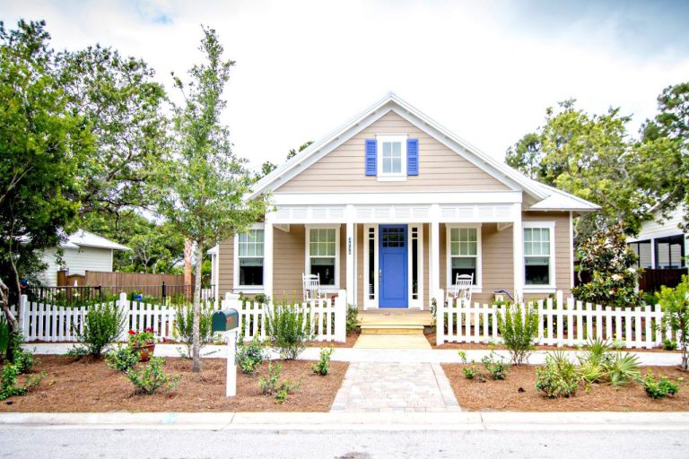 A charming beige house with a blue door and blue shutters, white trim, a front porch with chairs, a white picket fence, and a tidy garden, surrounded by trees and greenery on a sunny day.