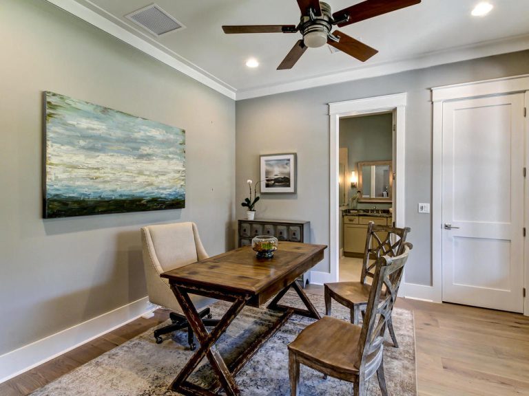 Home office with a wooden desk, three chairs, abstract art on gray walls, a ceiling fan, and a doorway leading to a bathroom with a large mirror and vanity. A potted plant sits on the desk.