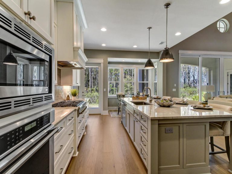 Modern kitchen with white cabinets, stainless steel appliances, a large island with sink, pendant lights, and wooden floors. Large windows and glass doors allow natural light and a view of trees outside.