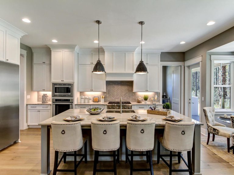 Modern kitchen with white cabinets, stainless steel appliances, island with marble countertop, four upholstered barstools, two pendant lights above, and a dining area visible in the background.