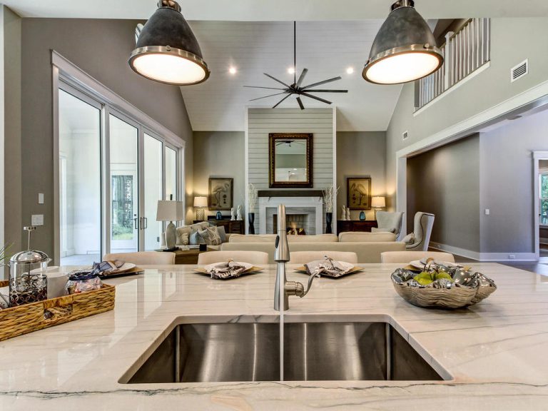 Modern kitchen island with a double sink in the foreground, overlooking an open living room with a fireplace, neutral decor, large windows, and high ceilings with hanging lights and a ceiling fan.