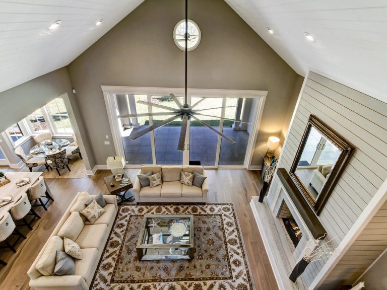 Spacious living room with vaulted ceiling, large windows, two beige sofas, glass coffee table on a patterned rug, and a fireplace with a mirror above it, viewed from above. Dining area visible on the left.