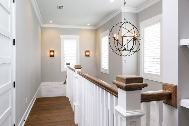 A modern hallway with light gray walls, white trim, wood flooring, white railing, and a spherical metal chandelier. Two wall sconces and windows with white blinds let in natural light.