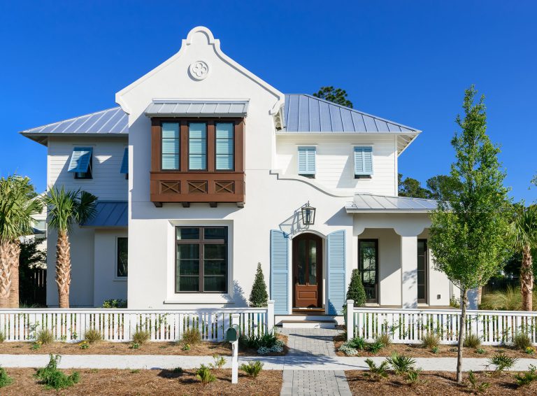 A white, two-story house with blue shutters and a metal roof. A white picket fence encloses the front yard with palm trees.
