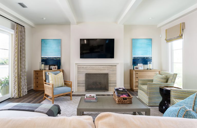 Bright living room with a mounted TV above a fireplace, two blue abstract paintings, light wood furniture, striped chairs, a gray coffee table with books, and large windows with curtains.