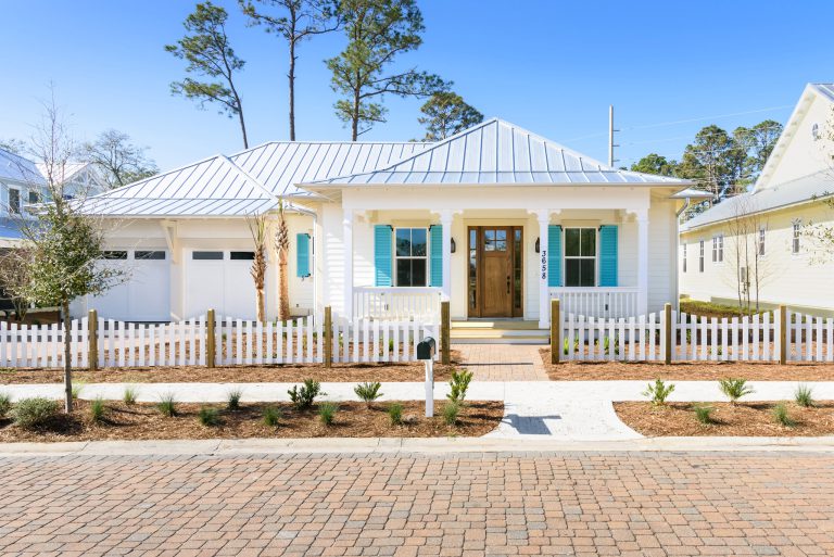 Single-story white house with a metal roof, turquoise shutters, and double wooden front doors, surrounded by a white picket fence, landscaped yard, and brick walkway under a clear blue sky.