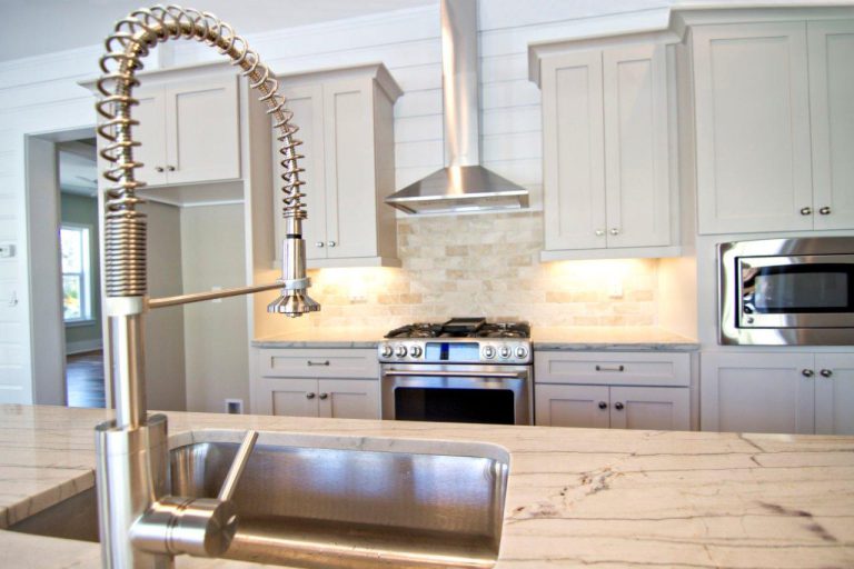 Modern kitchen with a marble countertop, stainless steel sink and spring-coil faucet in the foreground. In the background, there are light cabinets, a stainless steel stove with a hood, and built-in microwave.