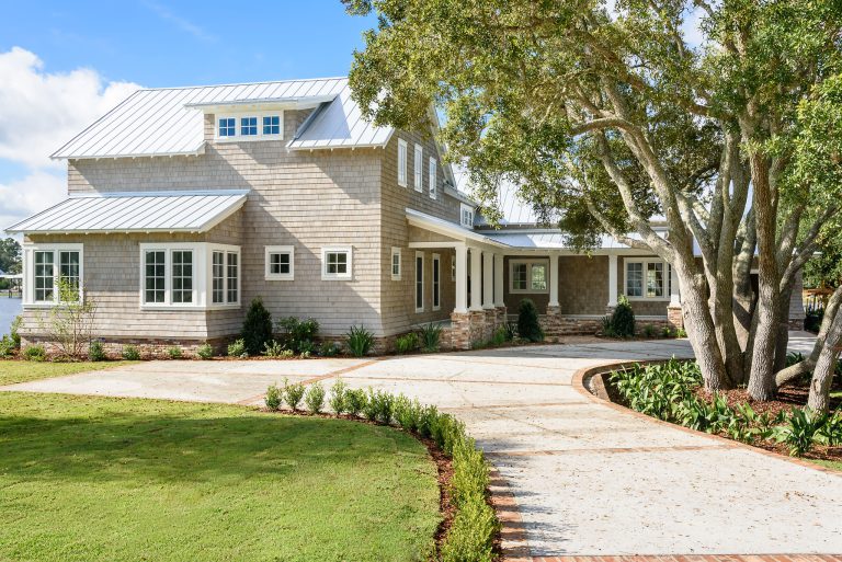 A modern two-story house with light gray shingles, large windows, and a metal roof, surrounded by a manicured lawn, landscaped garden beds, and tall trees with a curved stone driveway leading to the entrance.
