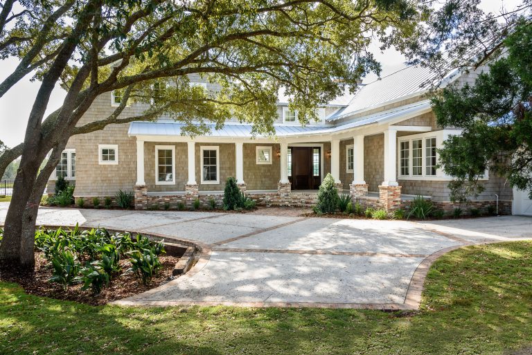 A large house with a wide front porch, brick and wood exterior, and manicured landscaping, shaded by tall trees. A curved driveway frames the garden beds and leads to the front door.