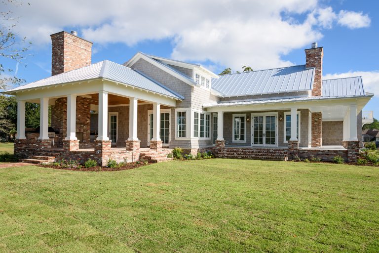 A modern farmhouse with a metal roof, large covered porch with white columns, brick accents, and multiple large windows, set on a neatly manicured lawn under a partly cloudy sky.