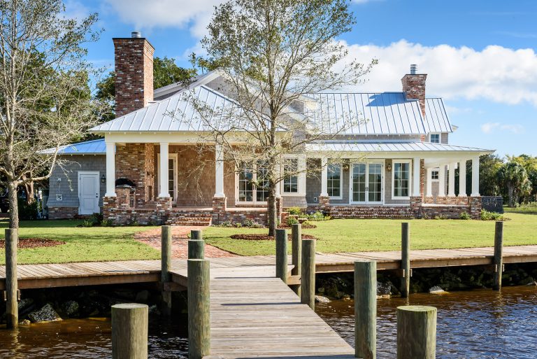 A large house with a metal roof, brick chimney, and wraparound porch stands behind a dock on a calm waterfront, with a grassy lawn, small trees, and blue sky in the background.