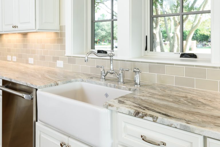 A modern kitchen countertop with a farmhouse sink, stainless steel faucet, beige subway tile backsplash, and white cabinetry beneath large windows showing green trees outside.