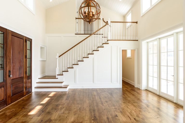 Bright, airy foyer with wooden floors, a white staircase with wooden handrails, large windows letting in natural light, glass-paned double doors, and a modern spherical chandelier hanging from the vaulted ceiling.