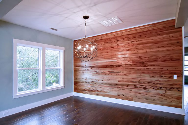 A modern, empty room with a wooden accent wall, dark wood flooring, large windows letting in natural light, and a stylish spherical chandelier hanging from the ceiling.
