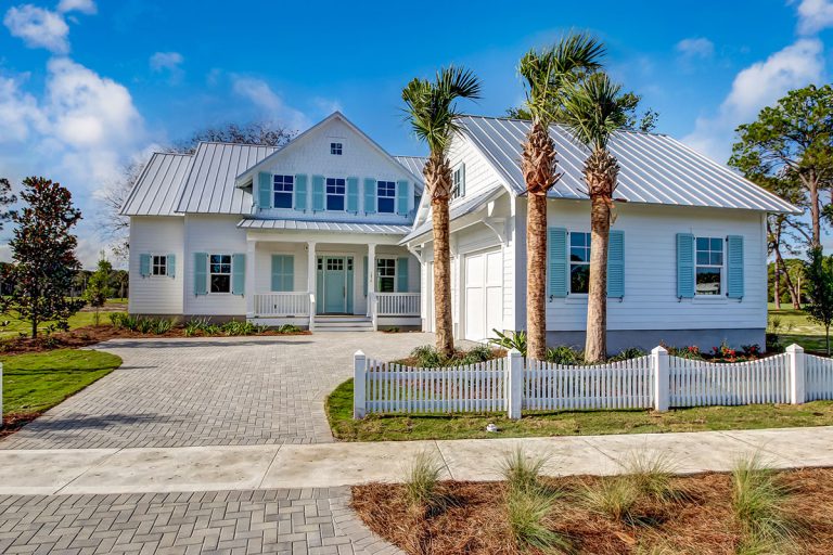A white, two-story house with a metal roof, blue shutters, palm trees, a white picket fence, and a paved driveway under a blue sky. Lush landscaping surrounds the home.