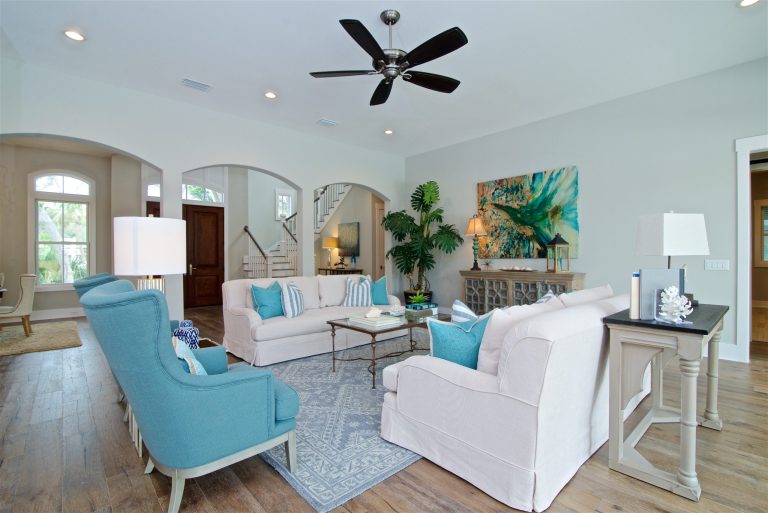 A bright, airy living room with white sofas, teal armchairs, blue and white pillows, a patterned rug, potted plants, coastal decor, wooden floors, and a ceiling fan. Natural light streams through large windows.
