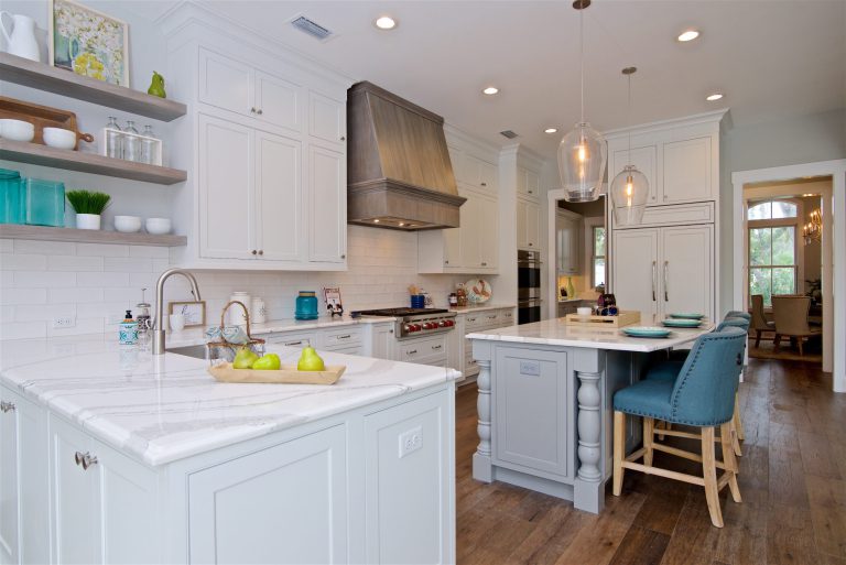 Modern kitchen with white cabinets, marble countertops, stainless steel appliances, a center island with teal barstools, open shelves with dishes, and decorative lighting fixtures above the island.