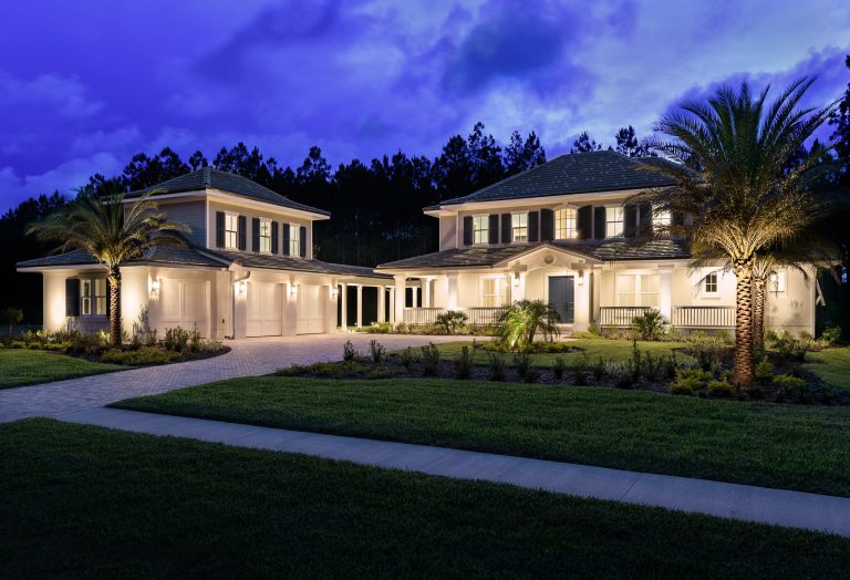 A large, elegant two-story house with bright exterior lights, manicured lawn, palm trees, and a driveway, photographed at dusk with a deep blue sky and trees in the background.