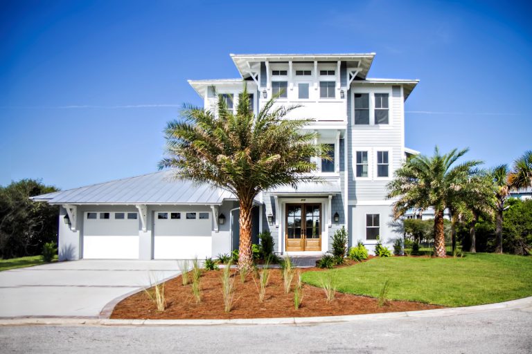 A modern three-story white house with large windows, a double garage, and a manicured lawn features palm trees and fresh landscaping under a clear blue sky.