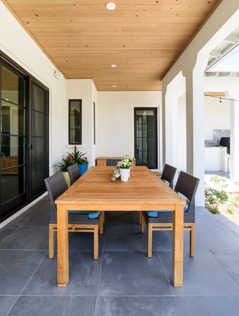 A modern covered patio features a wooden dining table with six chairs, a potted plant centerpiece, gray floor tiles, and white walls with large windows and doors.