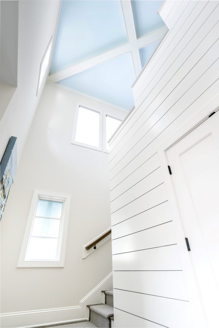 A bright, modern stairwell with white shiplap walls, a carpeted staircase, large windows letting in natural light, and a pale blue ceiling with decorative crossbeams.