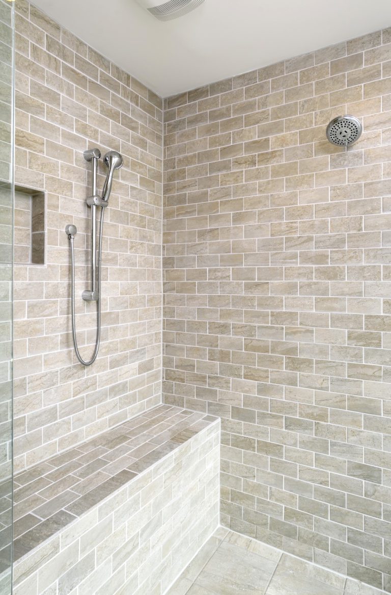 Walk-in shower with beige subway tile walls, a built-in tiled bench, a handheld showerhead with adjustable rail, and a fixed overhead showerhead. The space looks clean and modern.