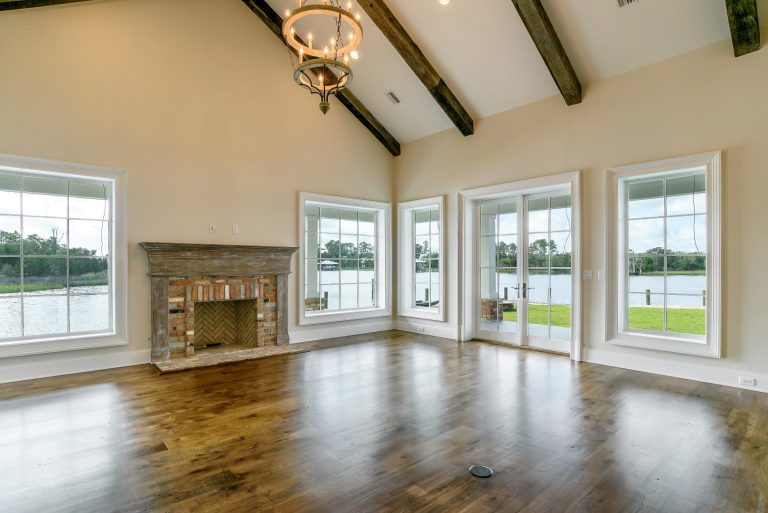 Spacious living room with hardwood floors, exposed wood ceiling beams, a brick fireplace, large windows, and glass doors showcasing a scenic lake view and green lawn outside.