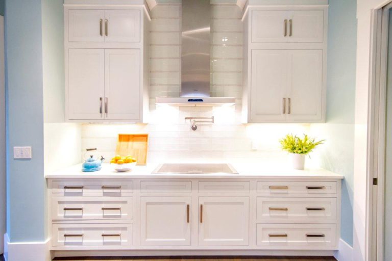 Bright, modern kitchen with white cabinets, stainless steel hood, white subway tile backsplash, and a white countertop. Items on the counter include a fruit bowl, cutting board, blue teapot, and a potted plant.
