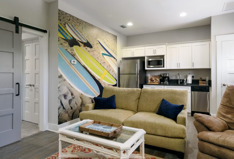 A modern living room with a yellow sofa, two navy pillows, a glass coffee table, and a large wall mural of surfboards on a sandy beach. The kitchen area features white cabinets and stainless steel appliances.