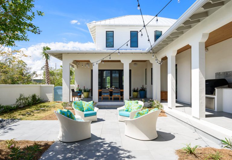 A modern white house with a covered patio, string lights, and four outdoor chairs with blue and green cushions arranged in a circle on a tiled patio. There is also an outdoor dining area and a built-in grill.