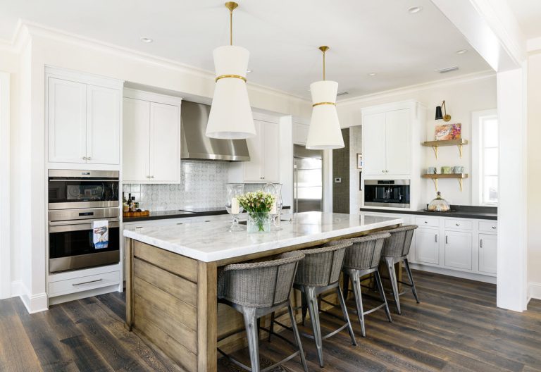 Modern kitchen with a large marble island, five wicker barstools, white cabinets, stainless steel appliances, pendant lights, and flowers in a vase on the island. Dark wood floors and open shelving add warmth to the space.